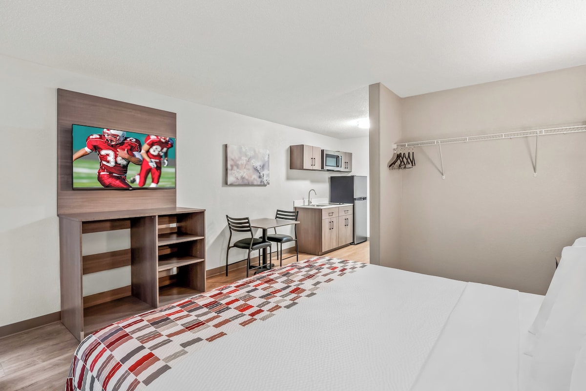 A hotel room with a bed, a TV mounted on a wooden shelf, a kitchenette, and a dining area with chairs.