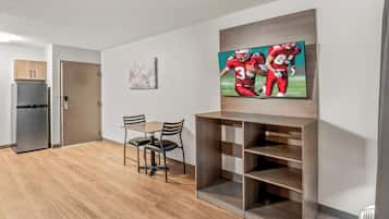 A modern living room with a flat-screen TV, wooden flooring, a dining table with chairs, and a built-in shelving unit.