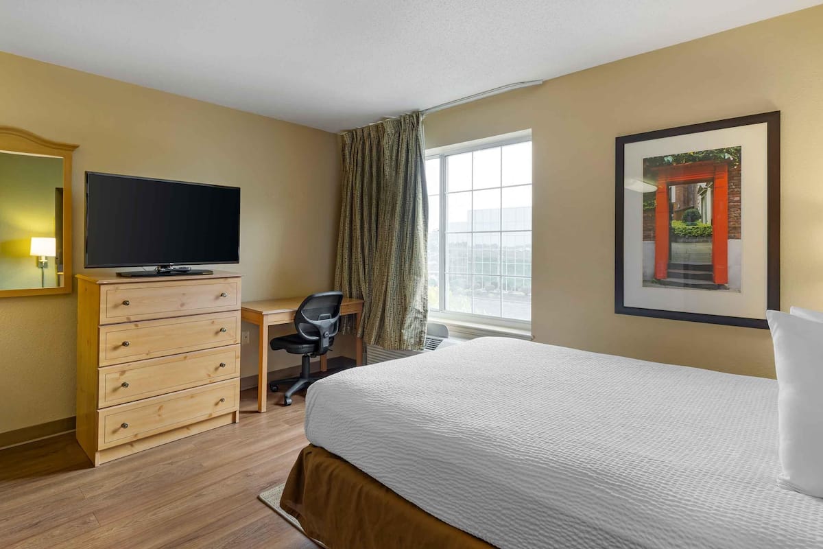 Habitación de hotel con cama, escritorio, televisor, cómoda, silla y ventana con cortinas.