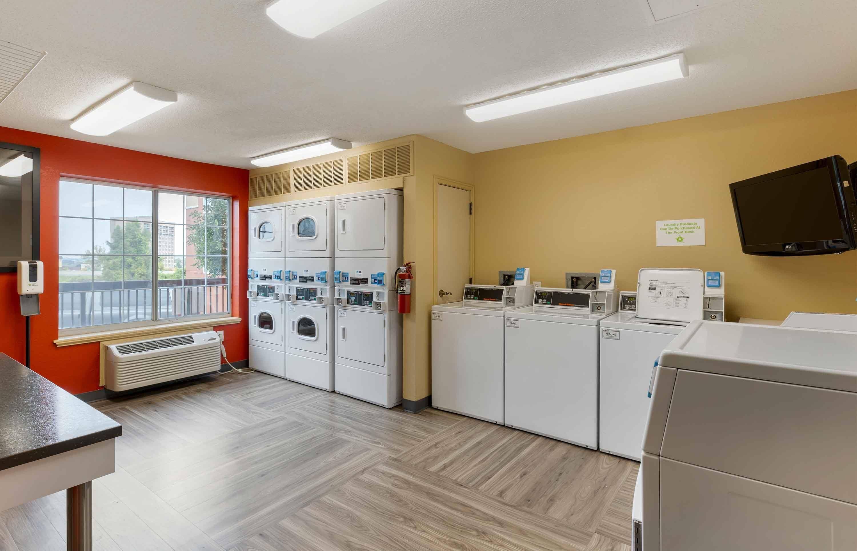 A laundry facility with multiple washing machines and dryers, a fire extinguisher, and a television mounted on the wall.