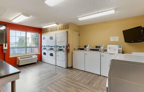 A laundry facility with multiple washing machines and dryers, a fire extinguisher, and a television mounted on the wall.
