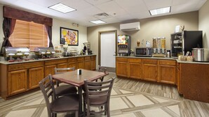 Una cocina de hotel con una mesa de comedor, sillas, microondas y diversos alimentos.
