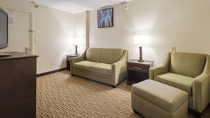 Habitación de hotel con sofá, dos mesitas de noche con lámparas, un pequeño reposapiés, un escritorio con una computadora portátil y un plano de evacuación contra incendios en la puerta.