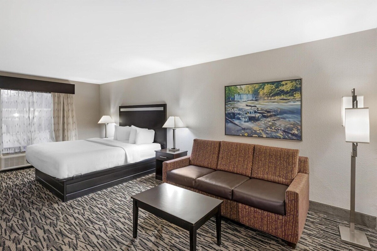 A hotel room with a bed, a sofa, a coffee table, a nightstand, a lamp, and a painting on the wall.
