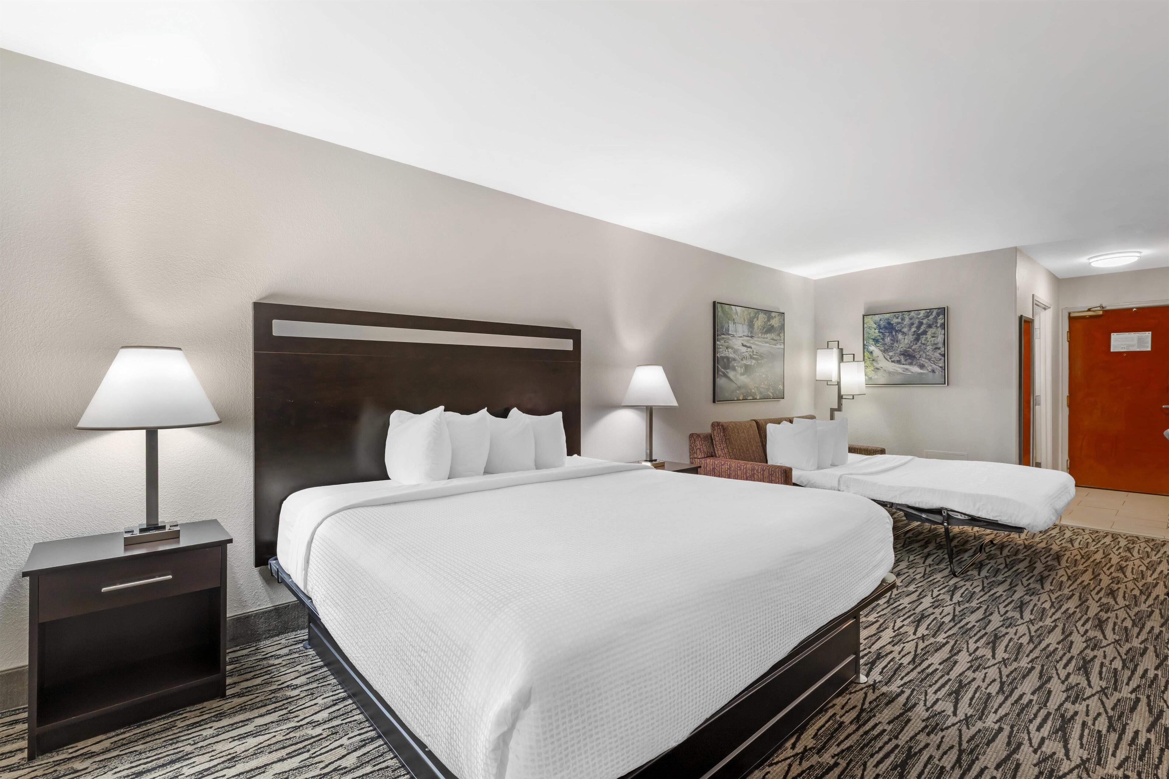 A hotel room with a large bed, two bedside tables, a nightstand, a patterned carpet, and a door.
