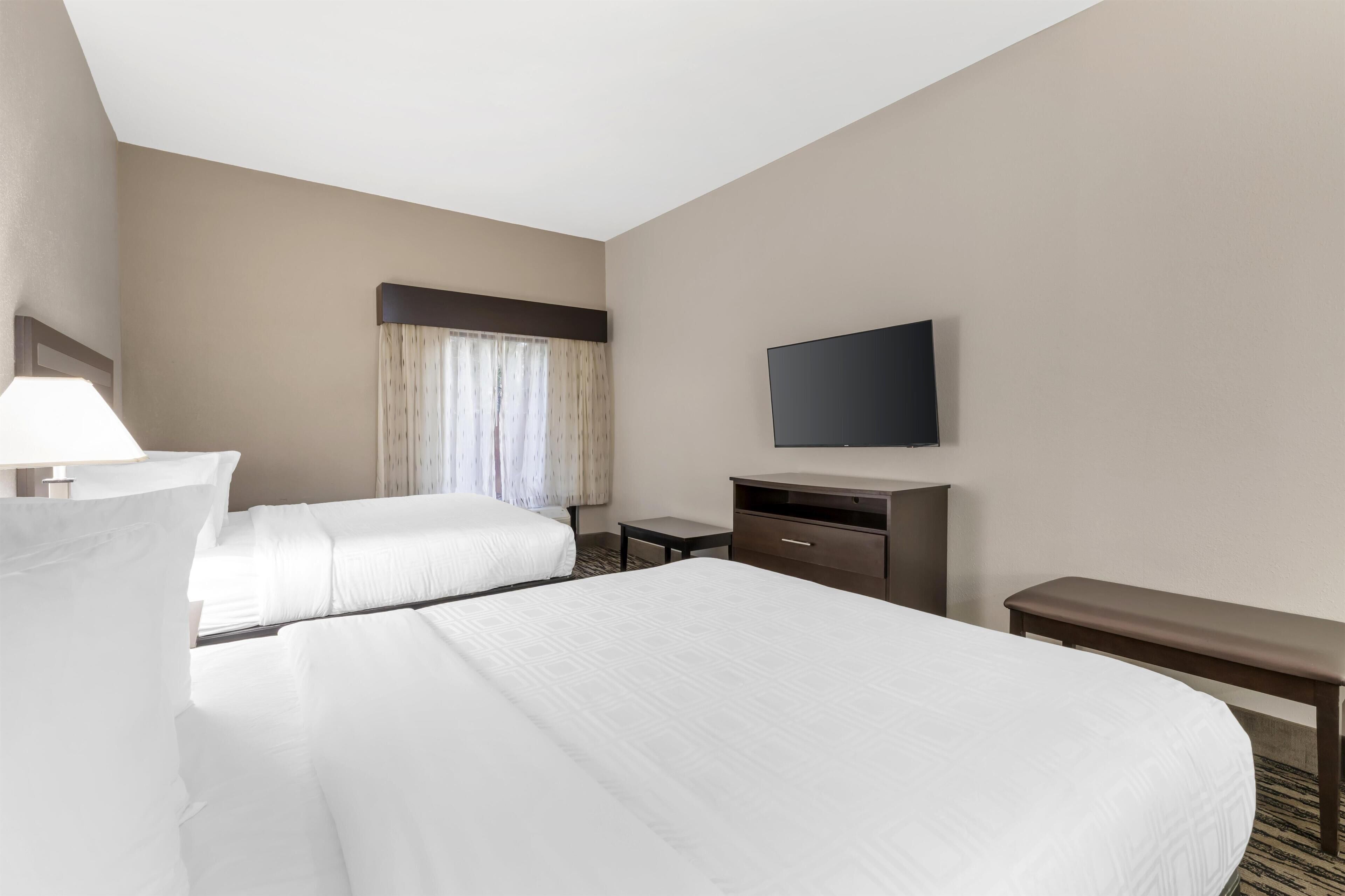 A hotel room with two beds, a flat-screen TV, a desk, and a bench.