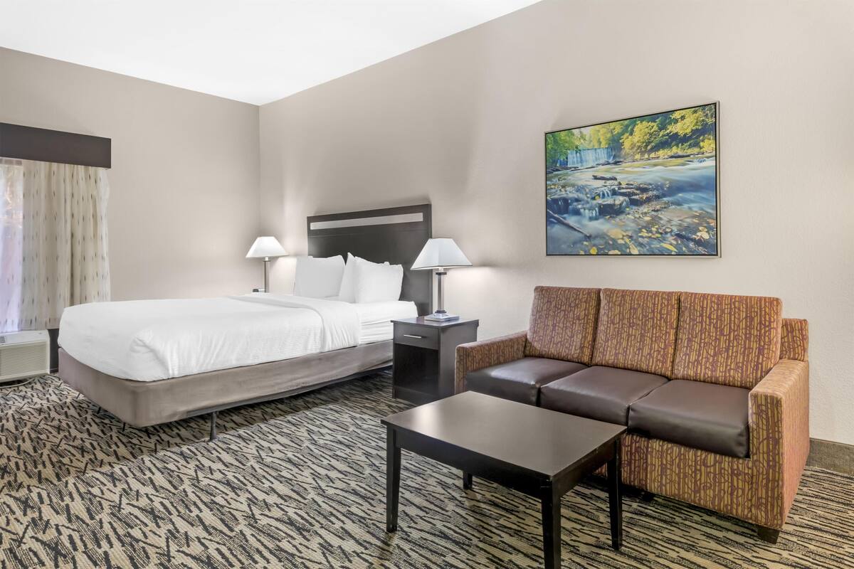 A hotel room with a bed, a sofa, a coffee table, a nightstand with lamps, and a painting of a waterfall.