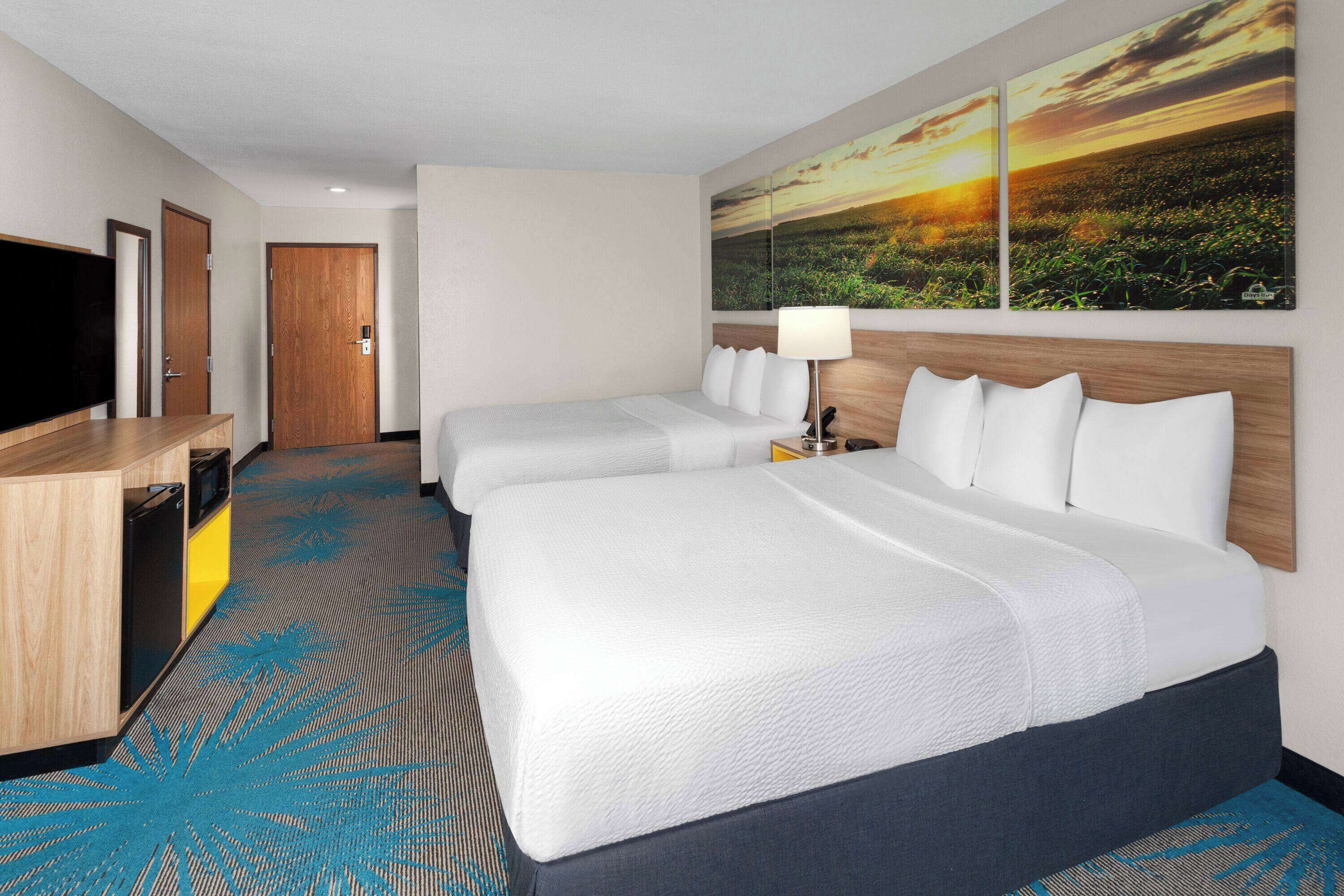 A hotel room with two beds, a TV, a desk, and a large painting of a sunset.