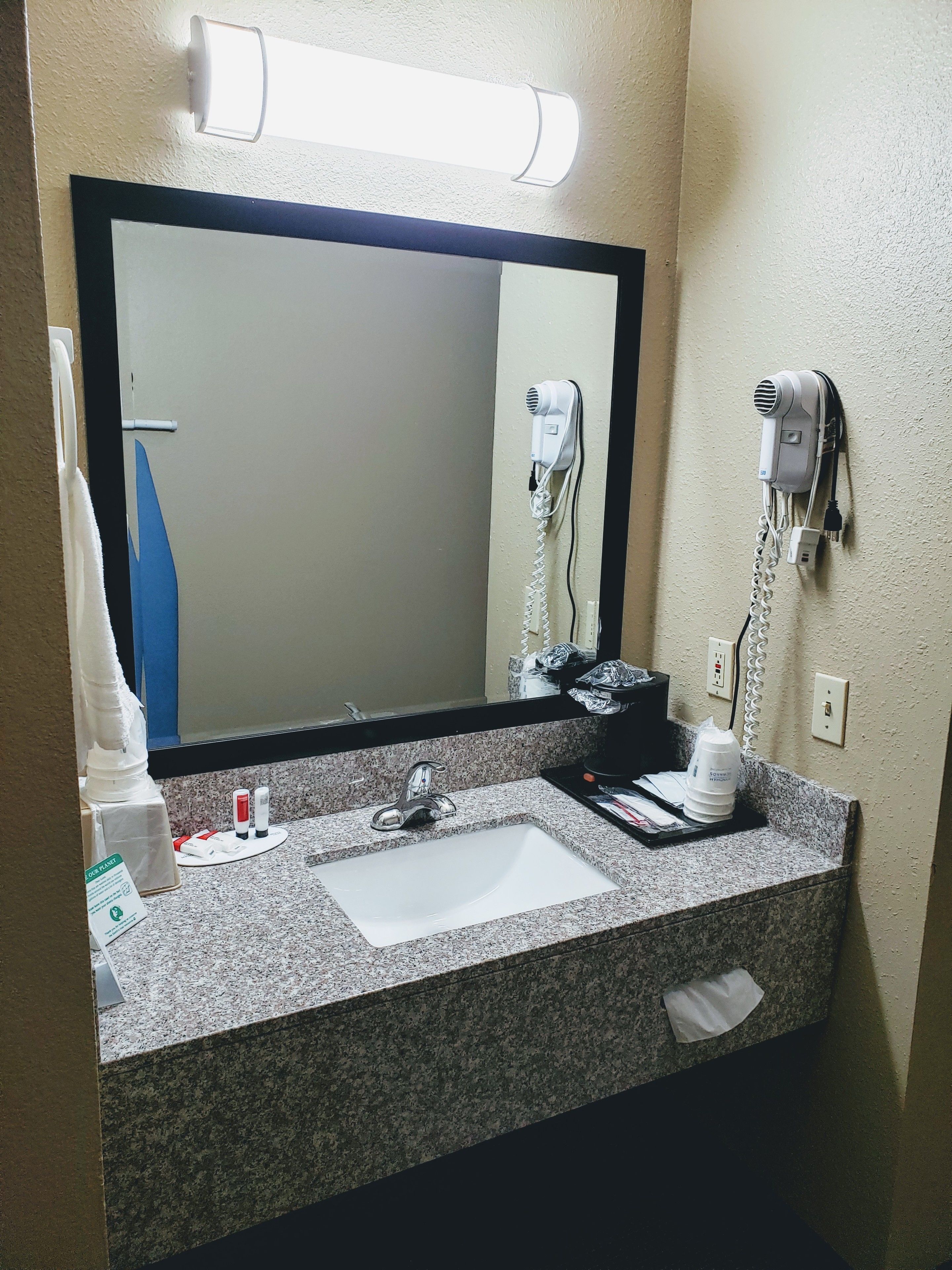 A hotel bathroom with a granite countertop, a sink, a mirror, a hairdryer, and a towel rack.
