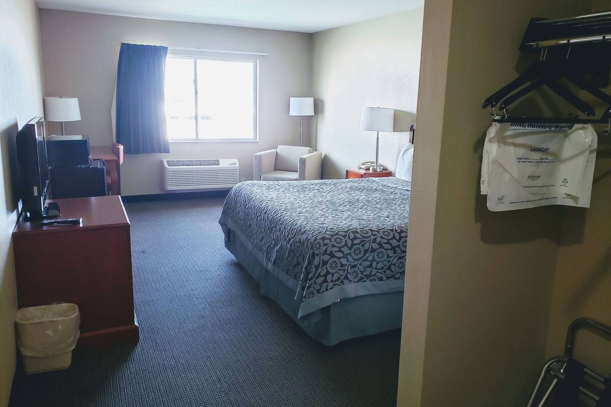 Habitación de hotel con cama, dos mesitas de noche, un escritorio con computadora, una silla, ventana con cortinas y perchero con una prenda colgada.