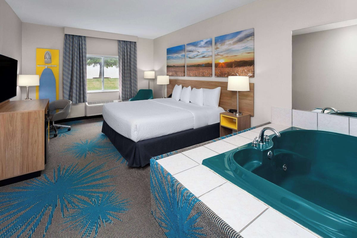Habitación de hotel con una cama grande, un escritorio, una silla, un televisor, una ventana con cortinas, un cuadro, una bañera y una alfombra con estampado de hojas.