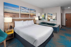 Quarto de hotel com uma cama grande, mesa de cabeceira com abajur, espelho, pia e uma cadeira.