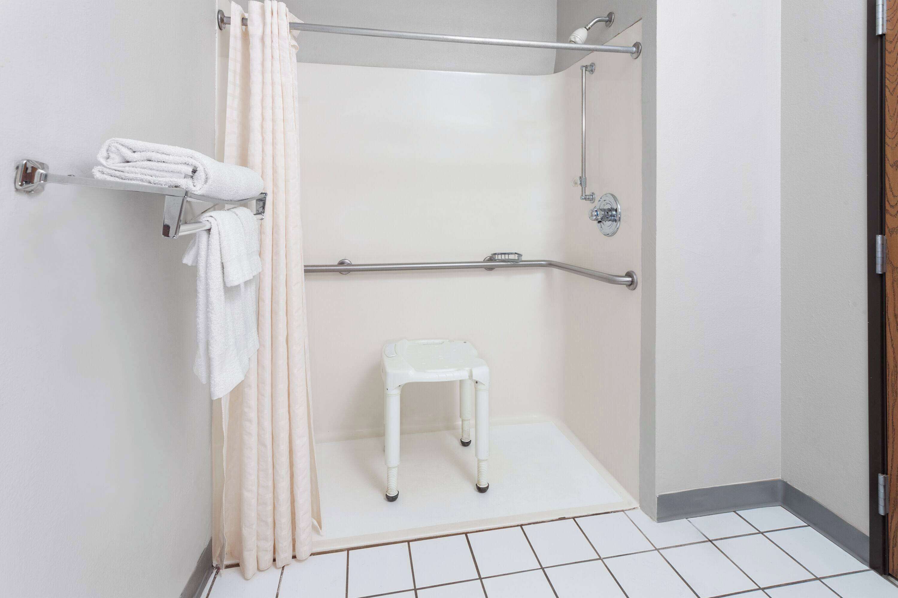 Un baño pequeño con azulejos blancos, zona de ducha, barra de apoyo y un taburete blanco.