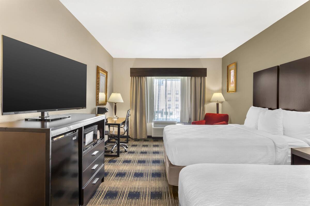 A hotel room with a flat-screen TV, a desk with a lamp, two beds, and a large window with curtains.