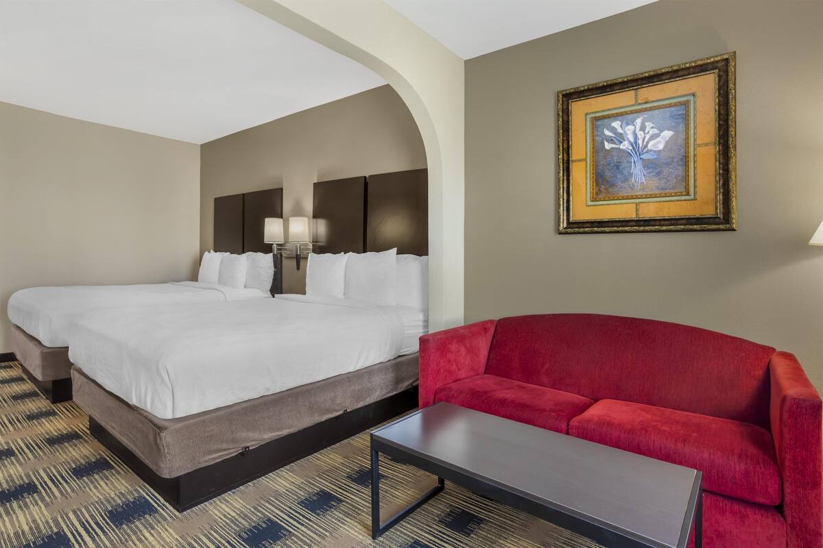 A hotel room with two beds, a red sofa, a small table, and a framed artwork on the wall.