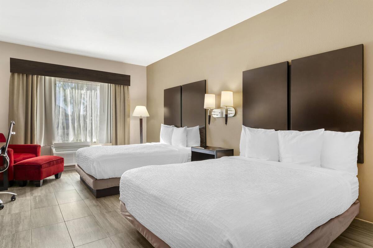 A hotel room with two beds, a red chair, a lamp, and a window with curtains.