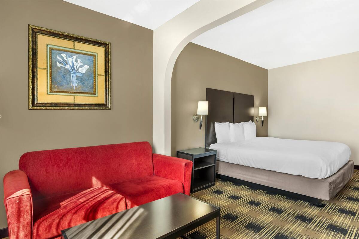 A hotel room with a red sofa, a bed with white linens, a framed artwork on the wall, and a patterned carpet.