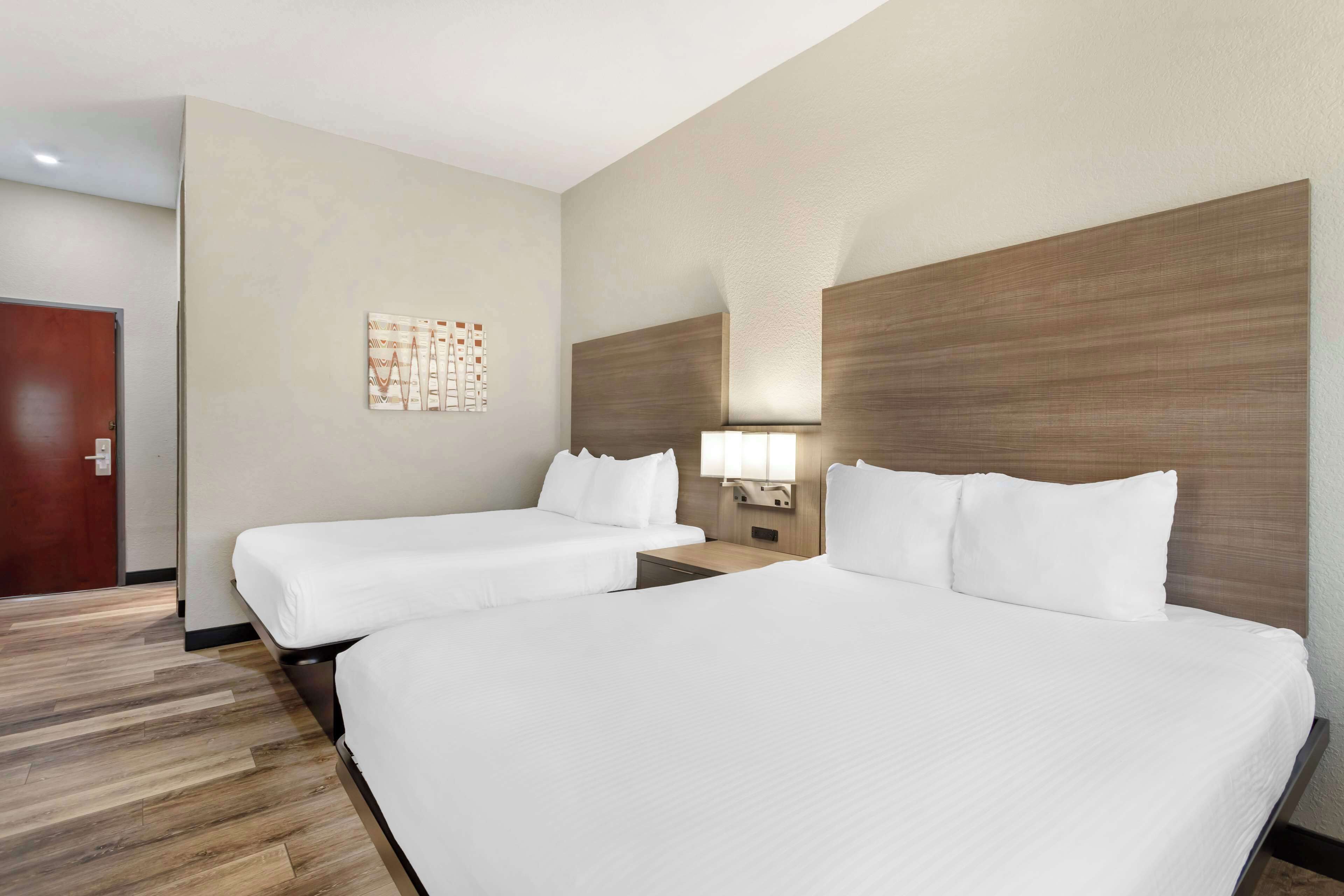 Une chambre d’hôtel avec deux lits, une tête de lit en bois, une table de chevet avec une lampe et une porte.