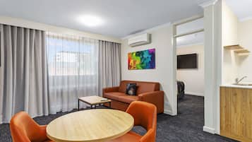 A hotel room with a round wooden table, orange leather chairs, a sofa, a TV, and an air conditioning unit.