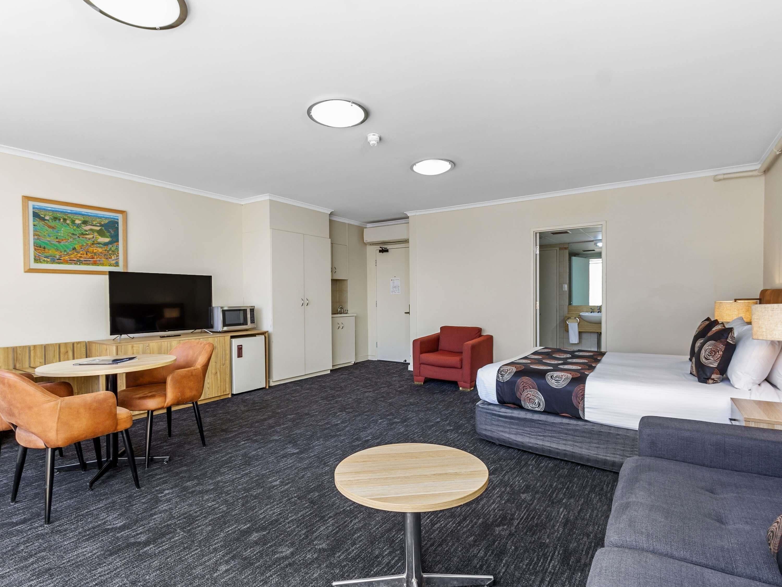 A hotel room with a bed, a sofa, a TV, a small kitchen area, and a dining table.