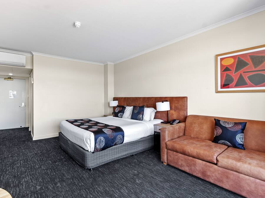 Une chambre d’hôtel avec un lit, un canapé en cuir marron, une table de chevet et un tableau abstrait accroché au mur.