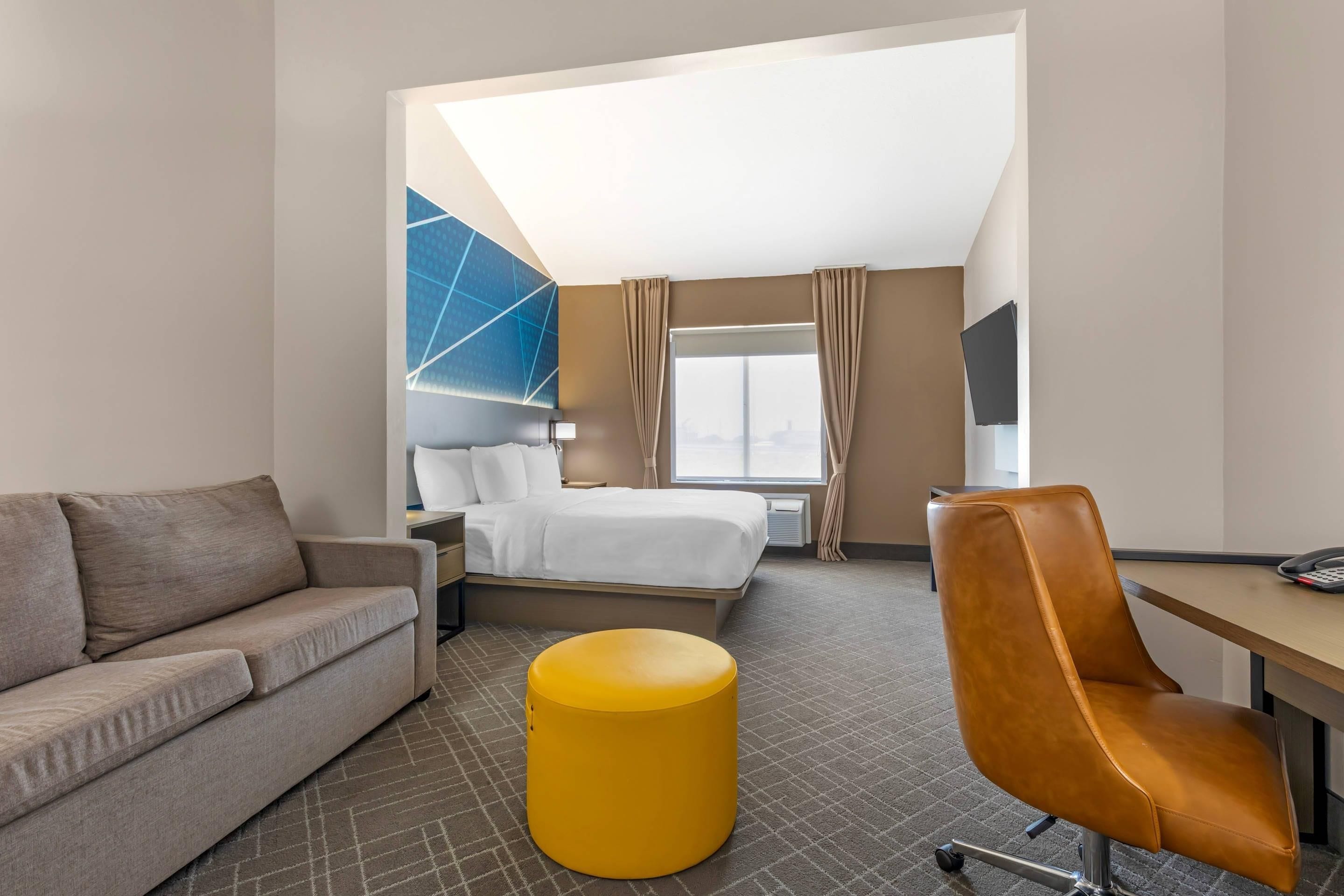 A hotel room with a bed, a sofa, a desk, a chair, a yellow ottoman, and a large window with curtains.