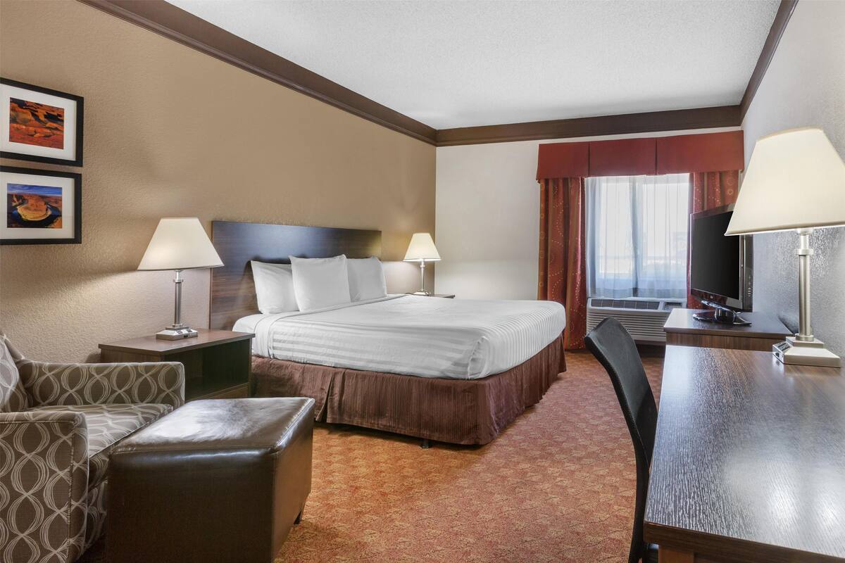 Ein Hotelzimmer mit einem Bett, zwei Nachttischen mit Lampen, einem Fernseher, einem Schreibtisch mit Stuhl und einem gemusterten Sessel.