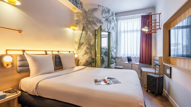 Quarto de hotel com uma cama grande, uma escrivaninha, um laptop e uma mala.