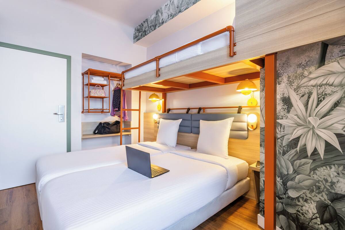 A double bed with a laptop, a wooden shelf with towels, and a wall-mounted lamp.