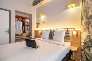 A modern hotel room with a neatly made bed, a laptop on the bed, a wooden headboard, and a built-in wardrobe.