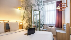 Quarto de hotel moderno com uma cama grande, um laptop sobre a cama, uma mesa pequena, uma cadeira e um espelho.