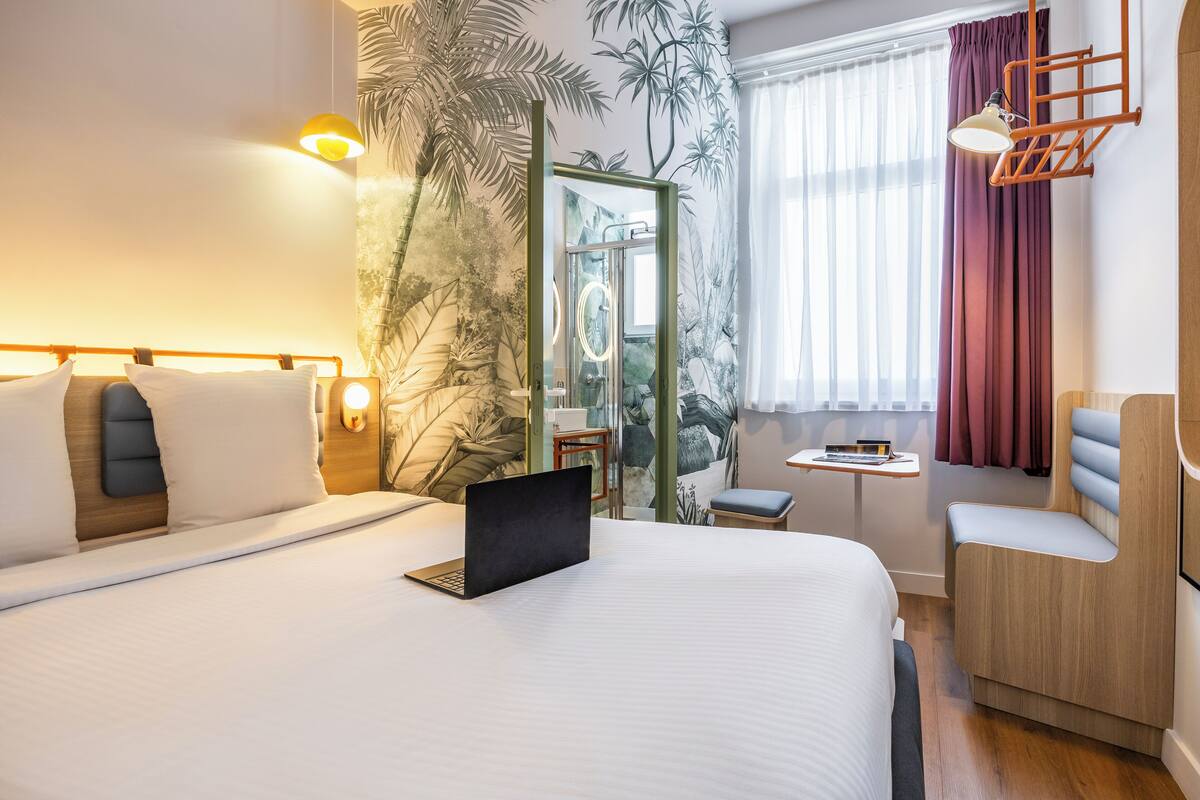 A modern hotel room with a large bed, a laptop on the bed, a small table, a chair, and a mirror.