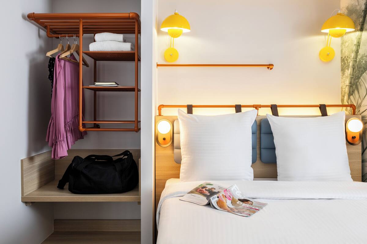 A hotel room with a wooden headboard, two bedside lamps, a bed with white linens, a magazine on the bed, a wooden shelf with a black bag and a pink garment, and a wooden wardrobe with hanging clothes and folded towels.