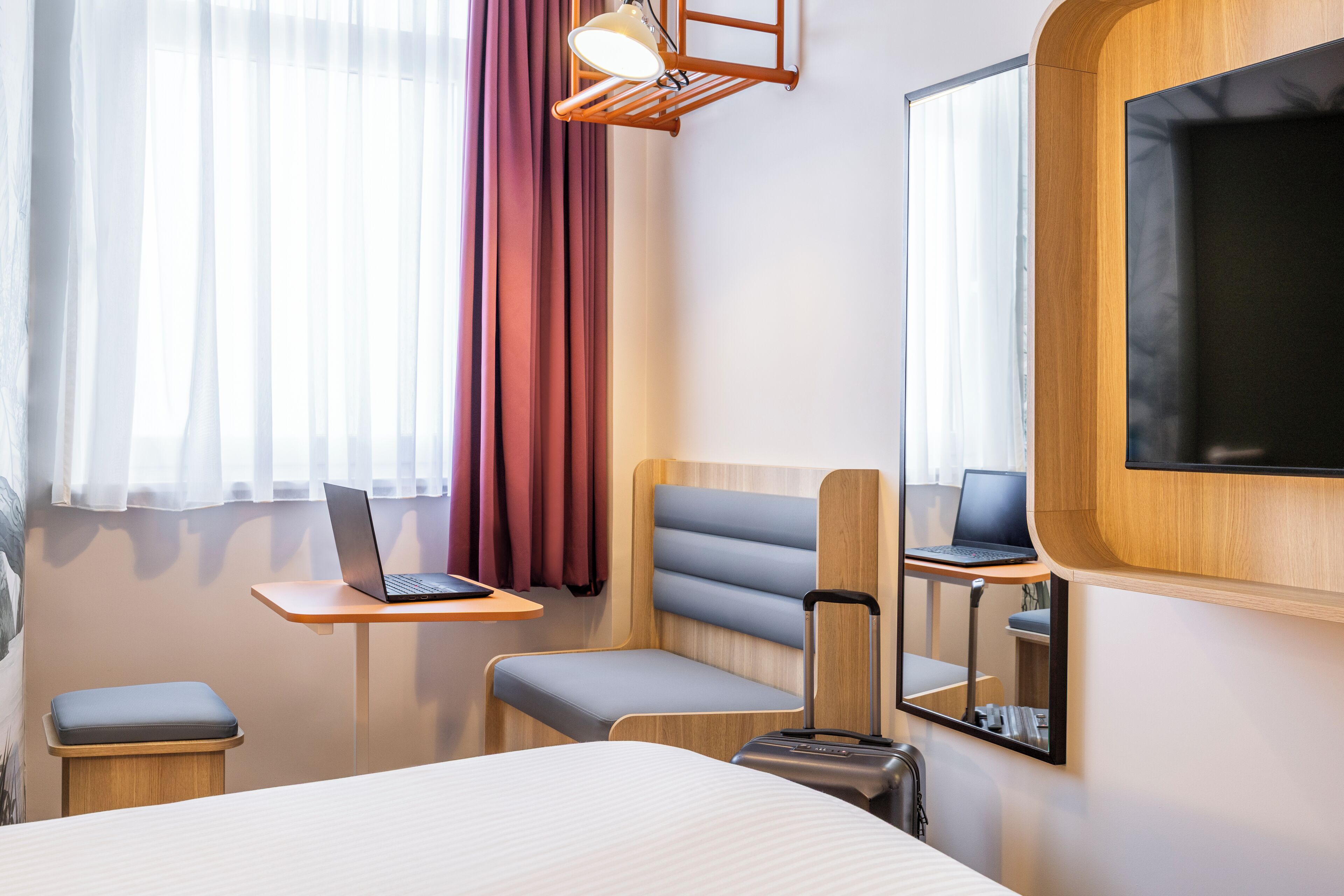 A hotel room with a bed, a desk with a laptop, a chair, a small table, a suitcase, a mirror, and a TV mounted on the wall.