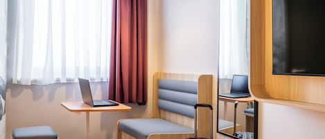 Habitación de hotel con cama, escritorio con ordenador portátil, silla, mesita pequeña, maleta, espejo y televisor en la pared.