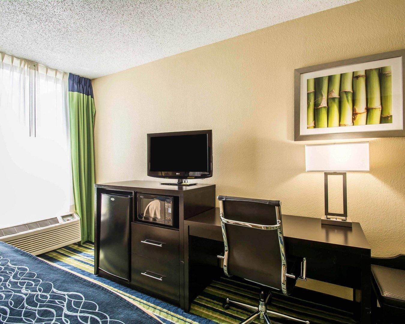 A hotel room with a flat-screen TV, a desk with a chair, a heater, and a painting on the wall.