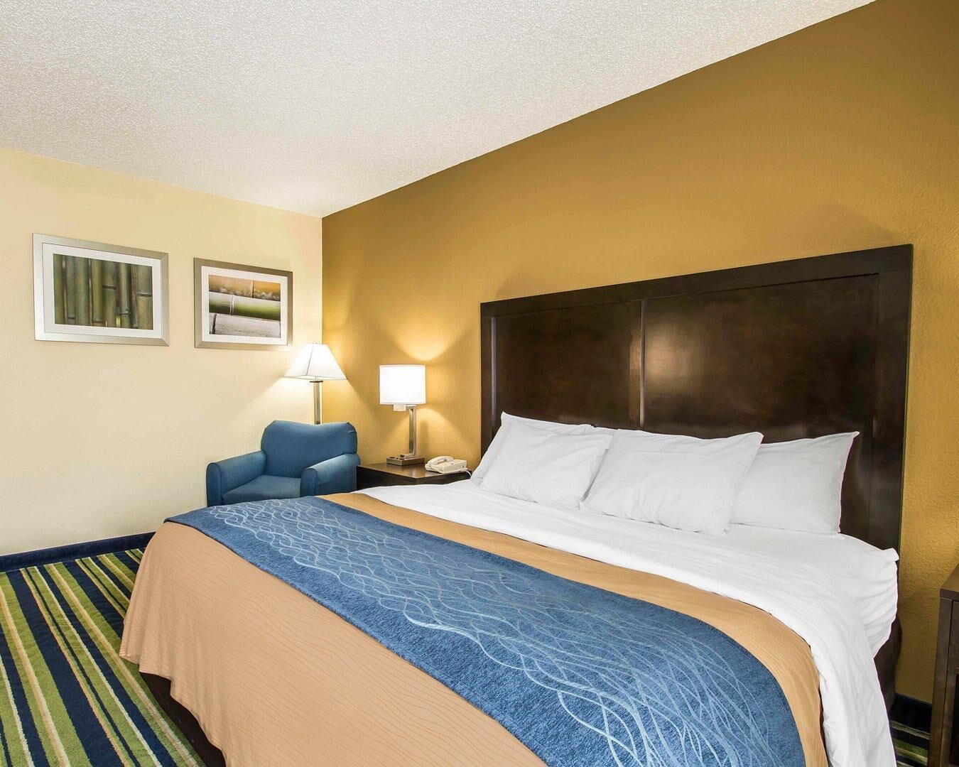 A hotel room with a bed, a chair, a nightstand, a lamp, and two framed pictures on the wall.