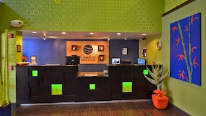 Zona de recepción de hotel con diseño moderno, que incluye un mostrador de recepción, un cartel de 'Comfort Inn & Suites' y una gran planta en maceta.