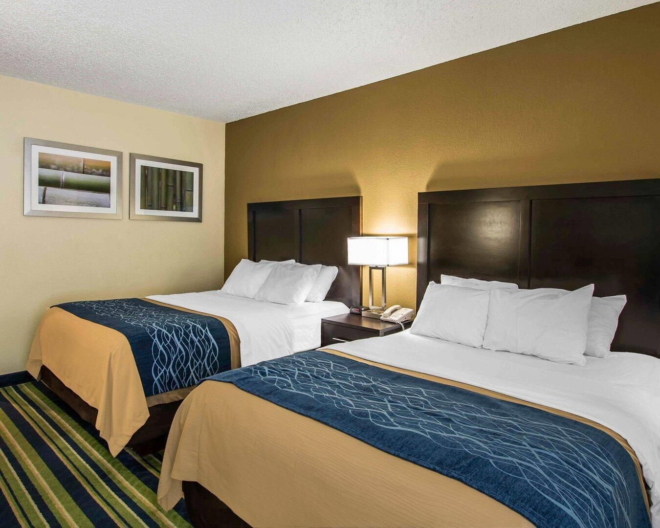 A hotel room with two beds, a nightstand, a lamp, and two framed pictures on the wall.