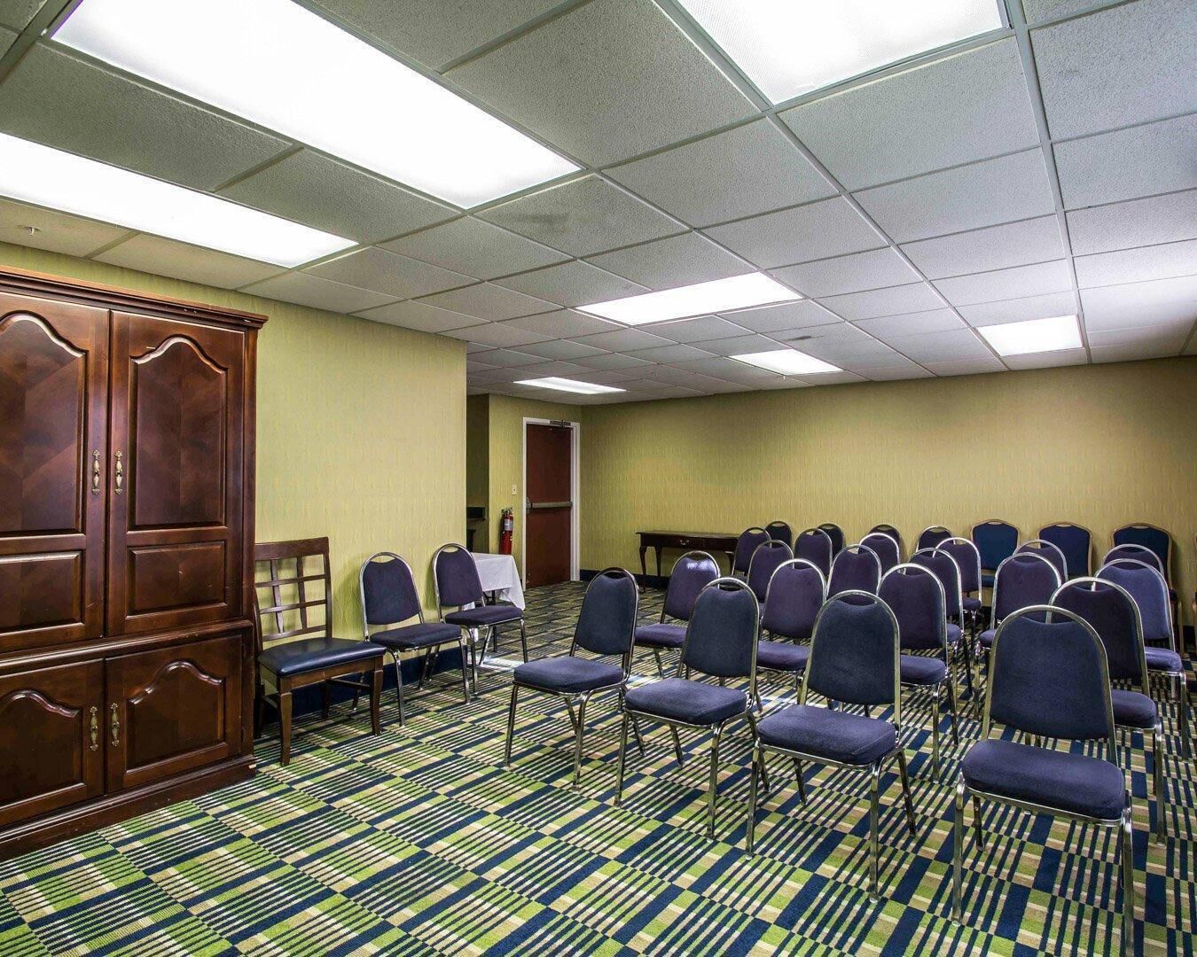 Una habitación de hotel con un mueble de madera grande, filas de sillas, una alfombra estampada y una puerta que da a otra habitación.
