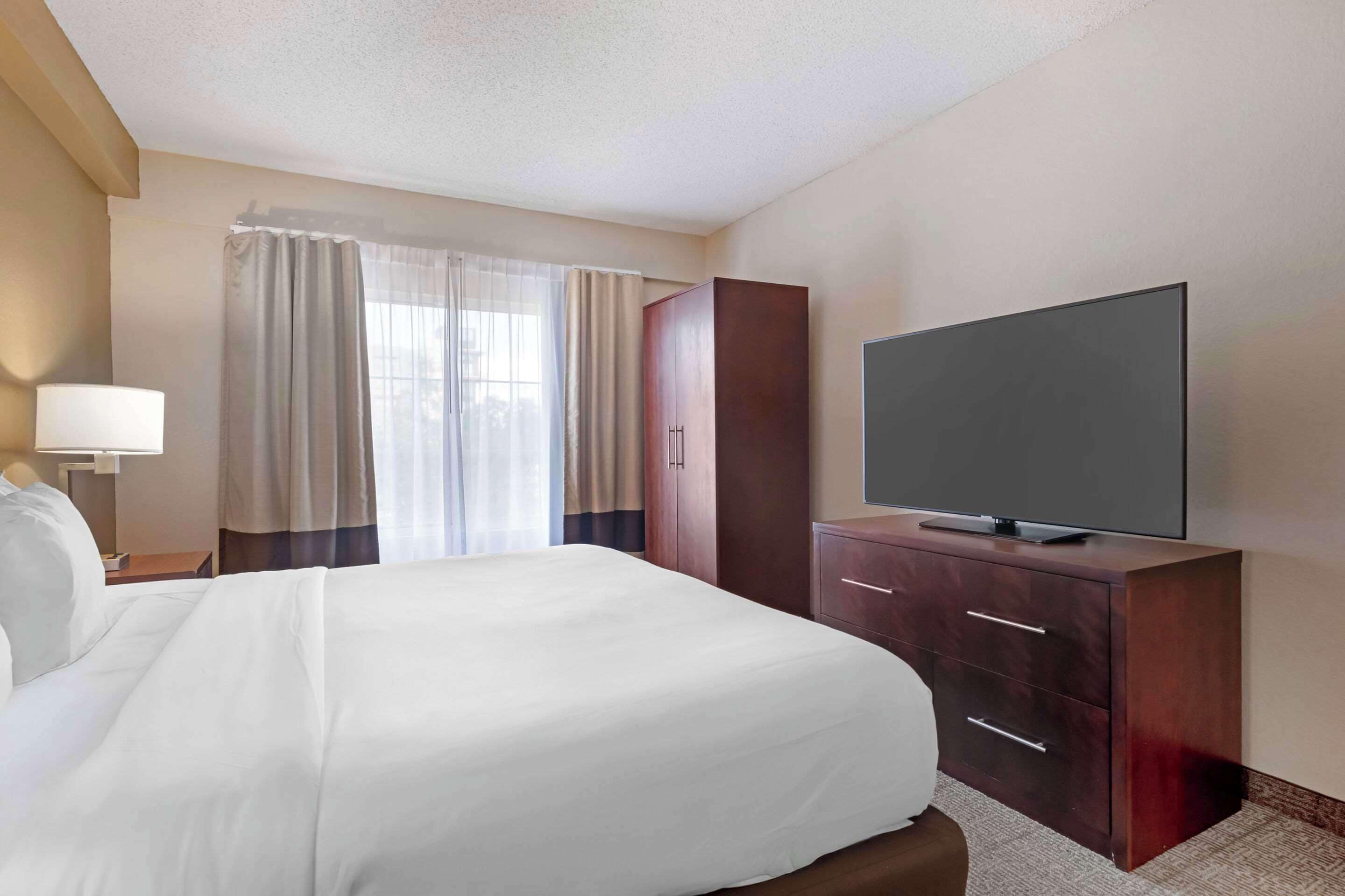 A hotel room with a large bed, a flat-screen TV on a wooden cabinet, a desk with a lamp, and a window with curtains.