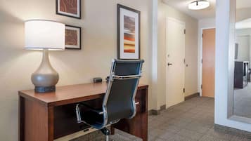 A hotel room with a desk, chair, lamp, framed artwork, and a door leading to another room.