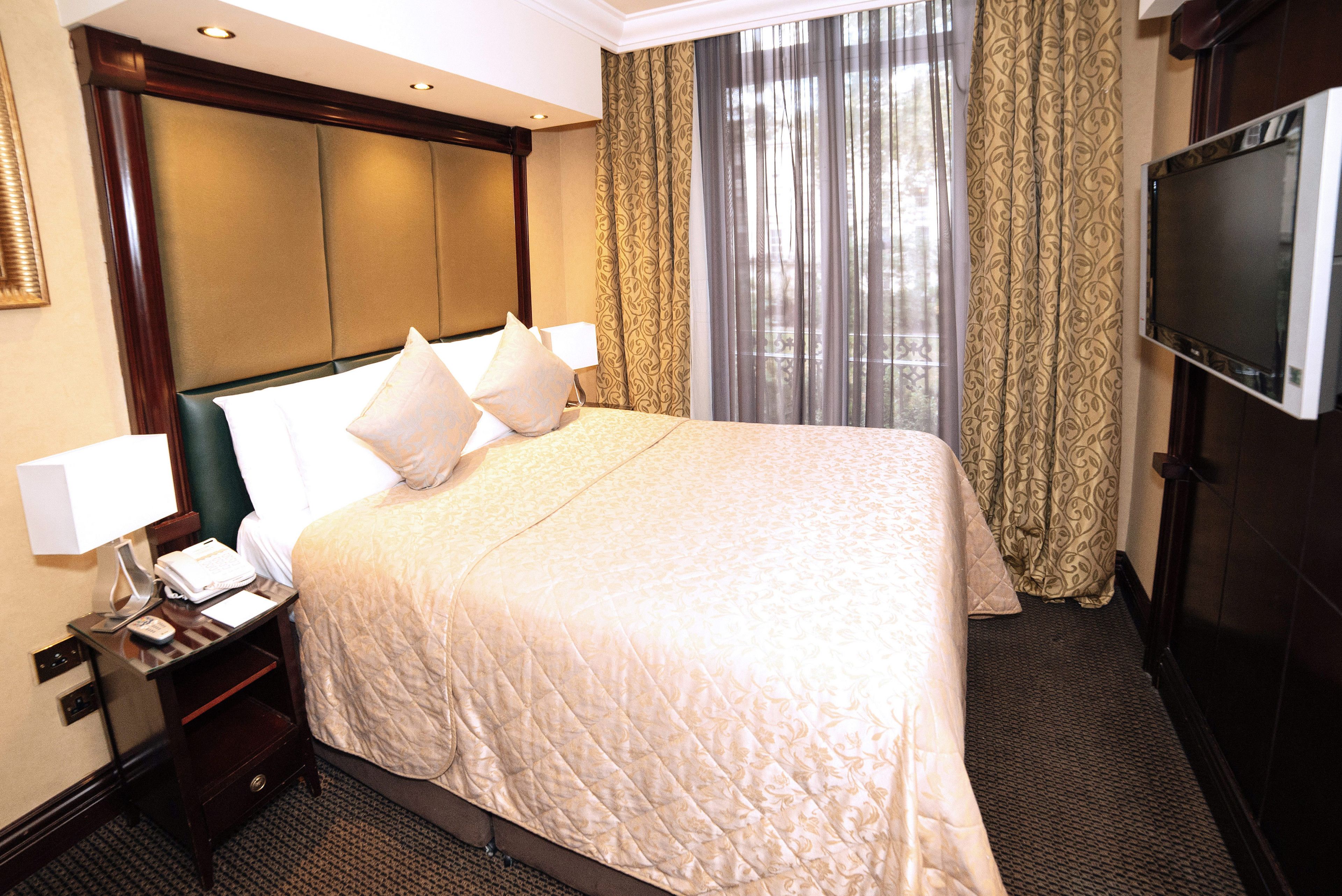 Een hotelkamer met een groot bed, een nachtkastje met een lamp, een televisie en een raam met gordijnen.