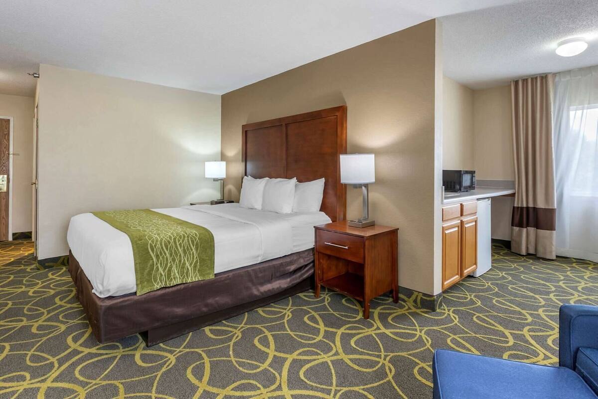 A hotel room with a bed, bedside table, lamp, and a kitchenette area.