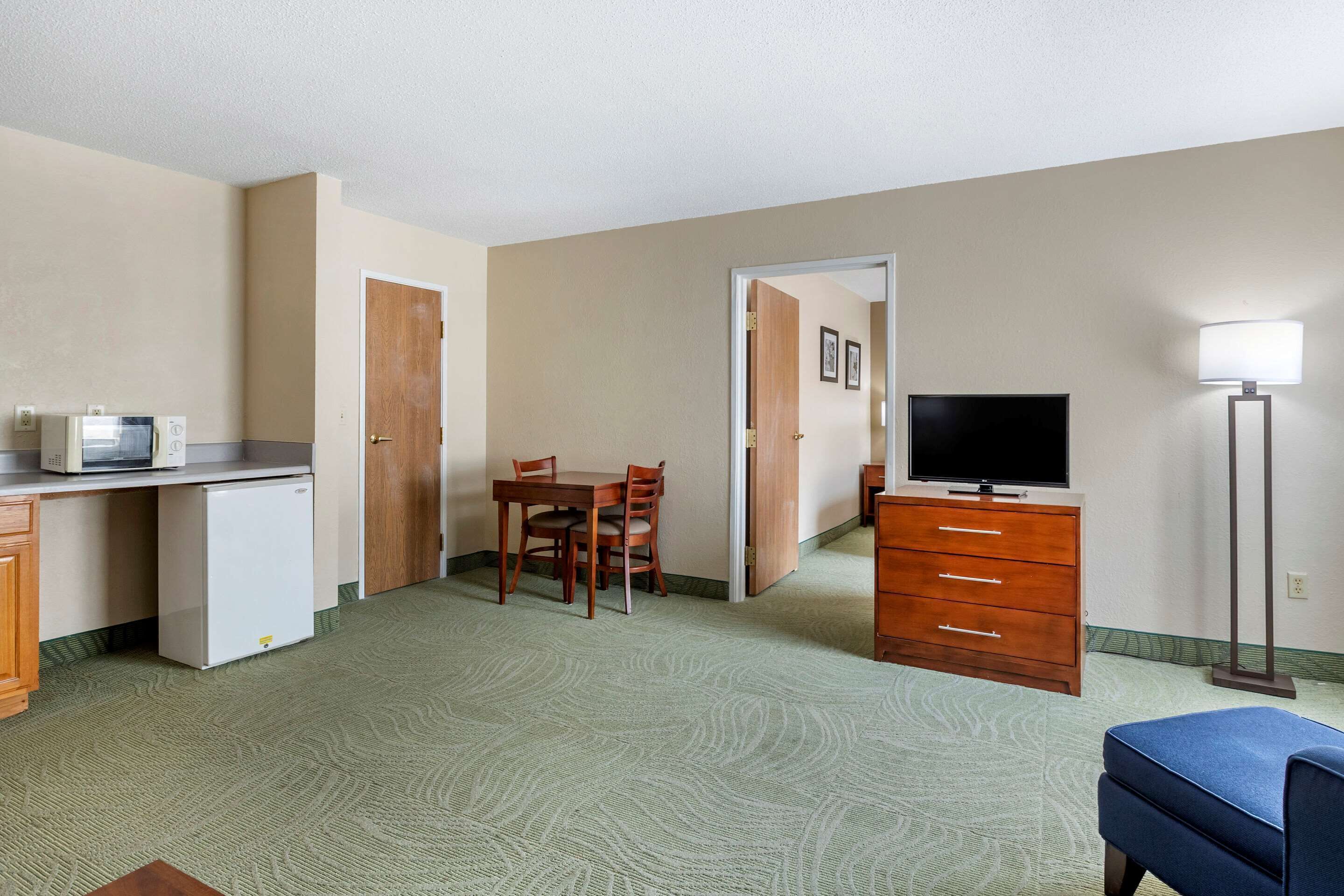 A compact hotel room with a kitchenette, a dining area, a TV, and a lamp.