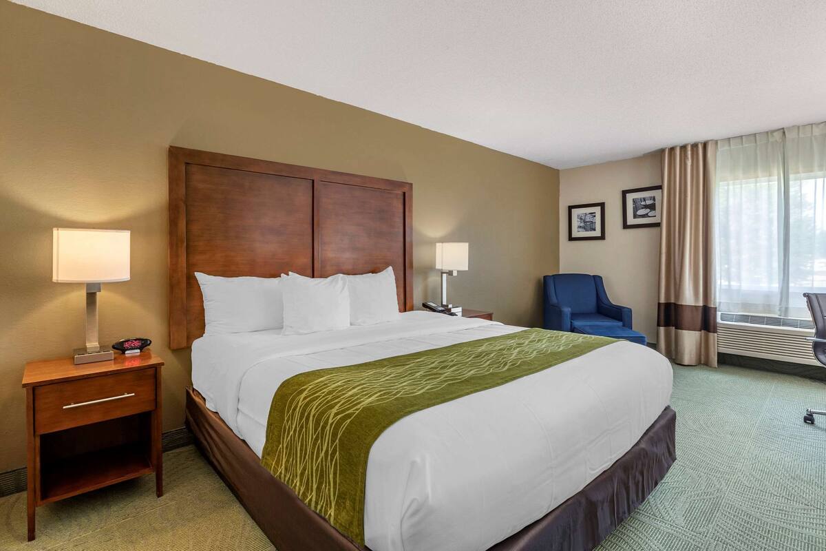 A hotel room with a large bed, a wooden headboard, two bedside tables with lamps, a blue chair, and a window with curtains.