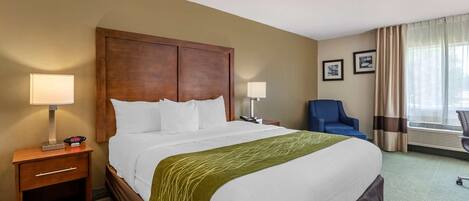 A hotel room with a large bed, a wooden headboard, two bedside tables with lamps, a blue chair, and a window with curtains.