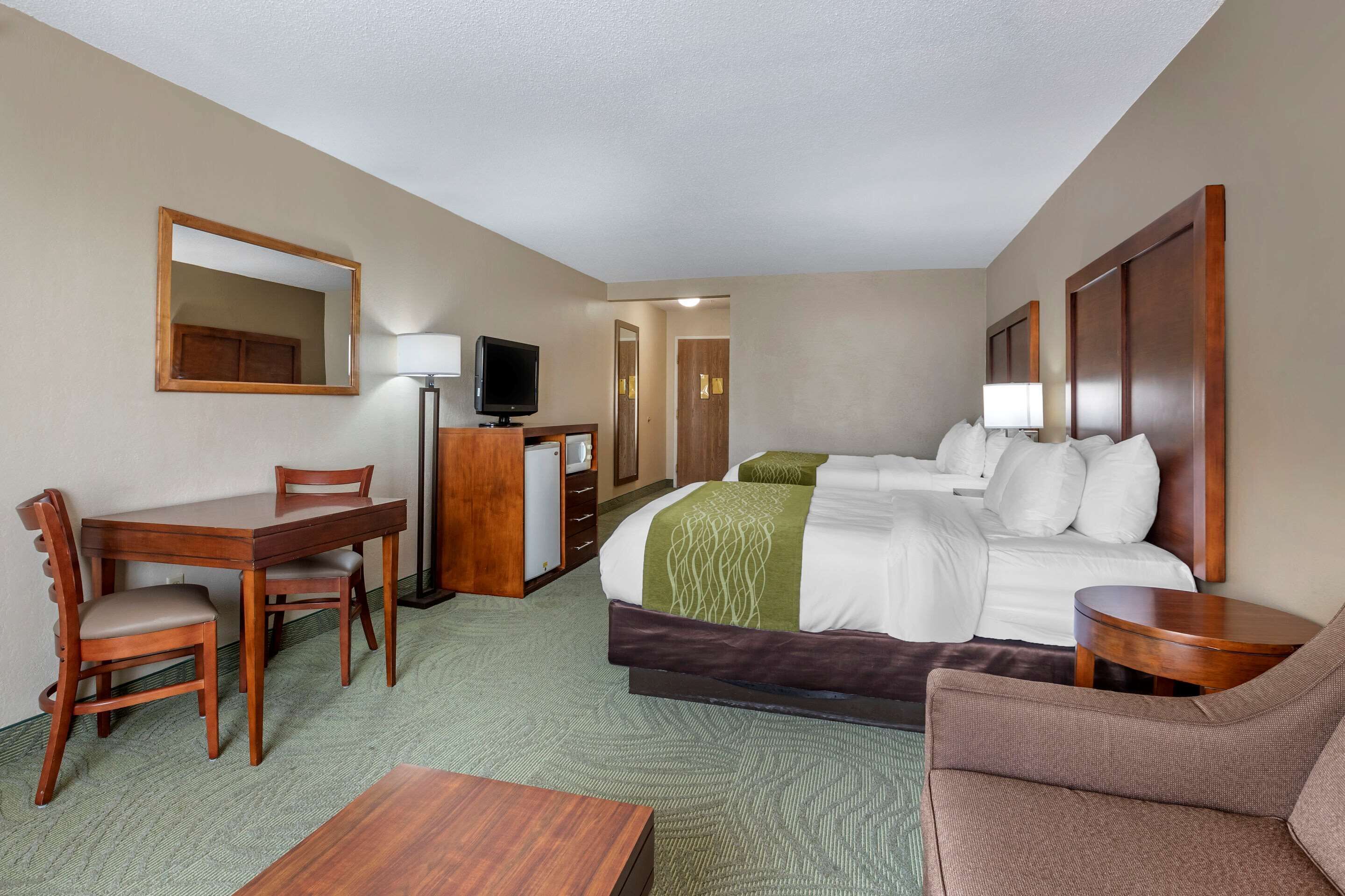 Quarto de hotel com cama, escrivaninha, cadeira, televisão e mesa pequena.