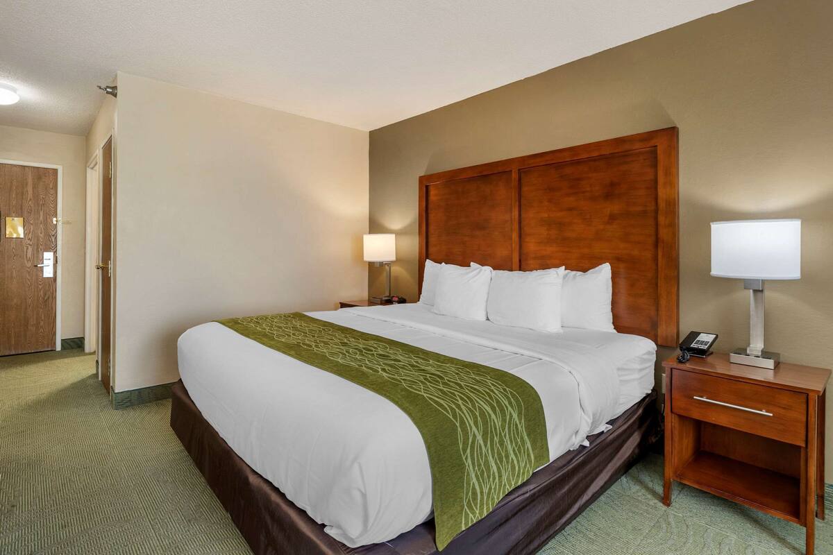 A hotel room with a large bed, wooden headboard, bedside lamps, a nightstand, and a carpeted floor.