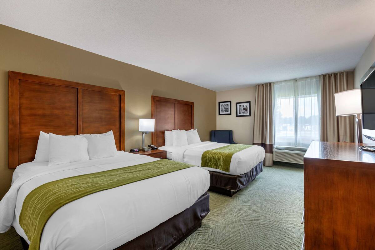 A hotel room with two beds, a desk, a chair, a lamp, and a TV.